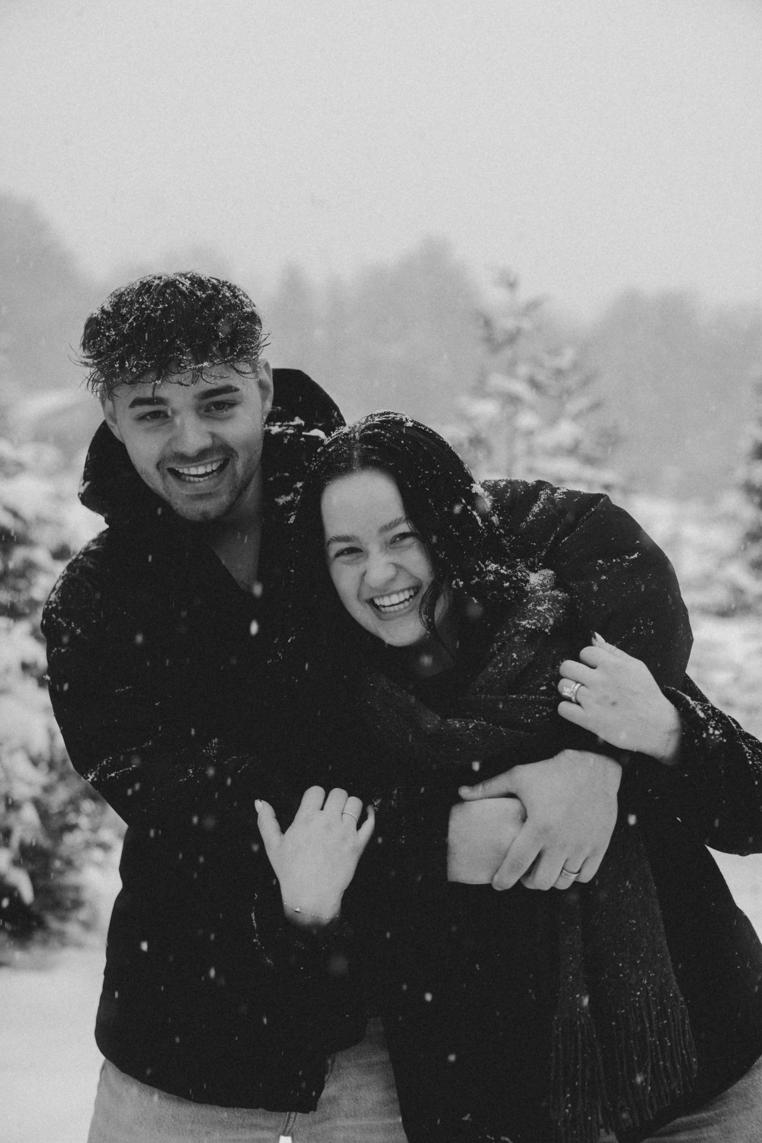 A couple laughs and embraces while enjoying a snowy day among snow-covered evergreen trees.