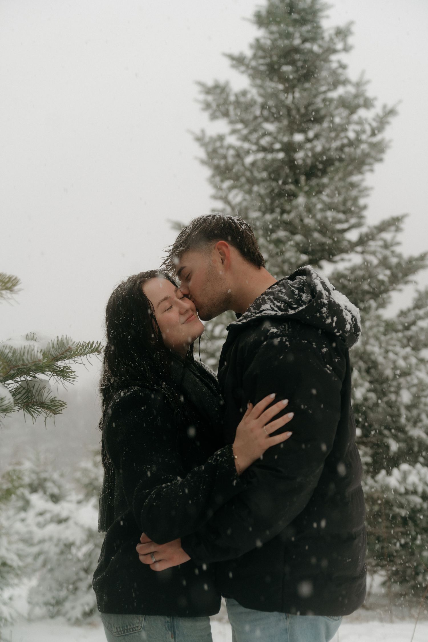 Romantic couple embracing in snowy forest with evergreen trees in background.