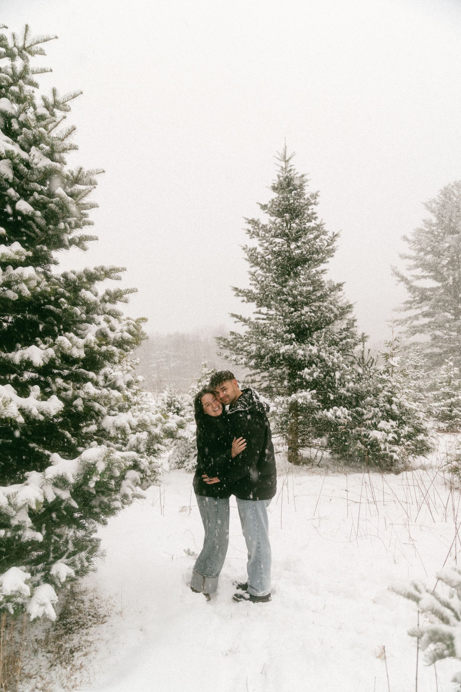 A couple shares an intimate moment in a snowy winter landscape surrounded by evergreen trees.