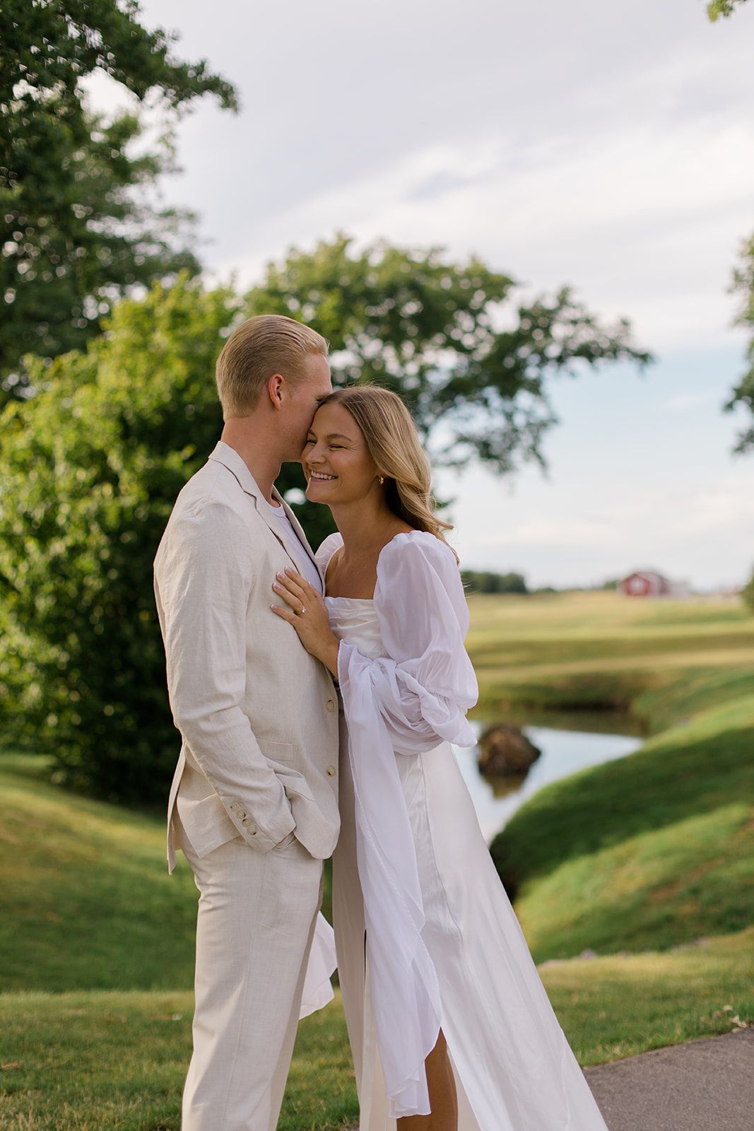 A romantic couple in white outfits embrace on a lush green hillside with a winding path and trees in the background.