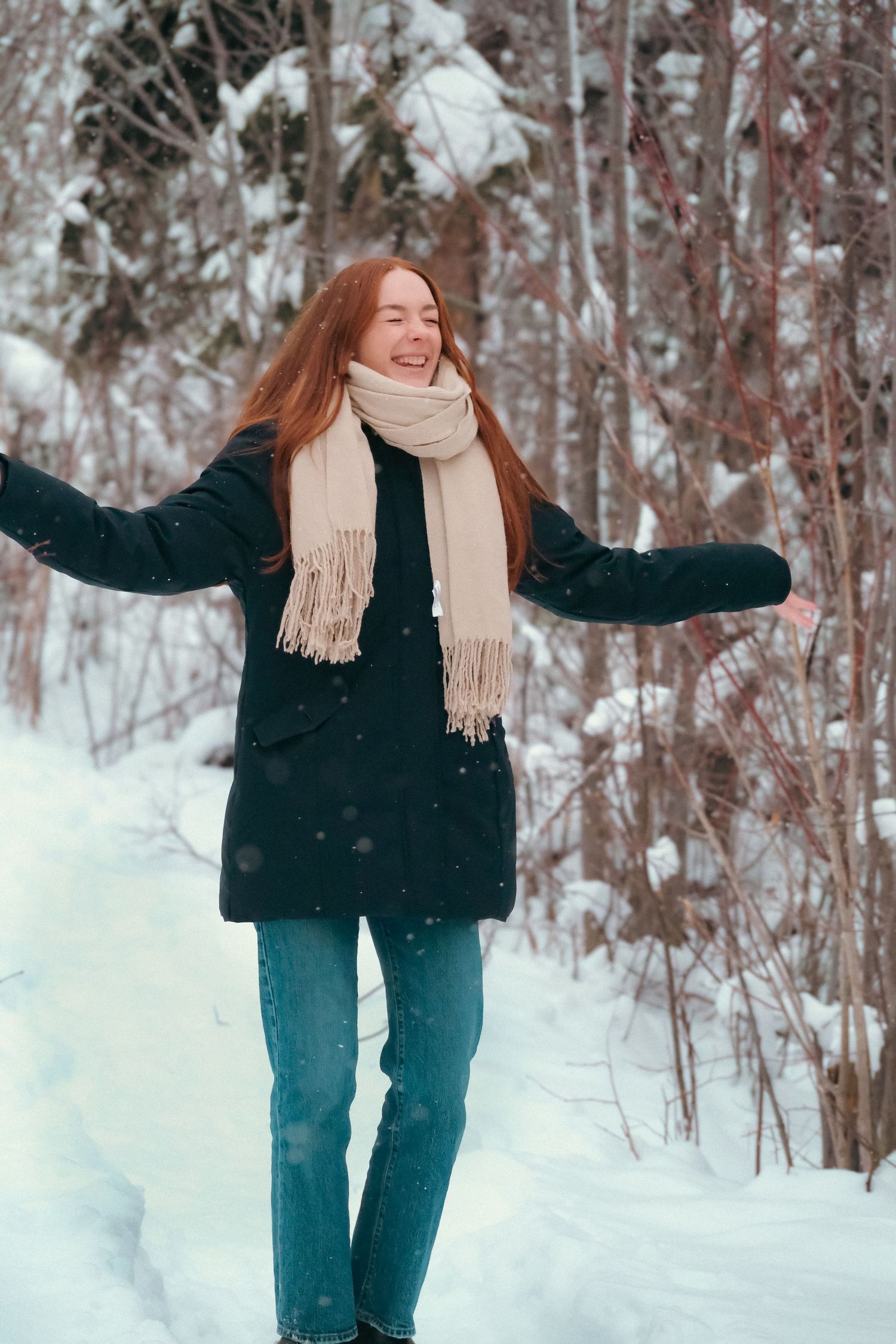 Person in black coat and beige scarf playing joyfully in snowy winter landscape.