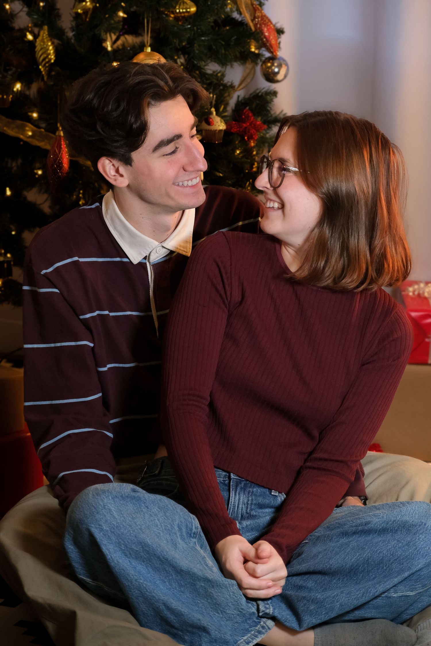 Couple in burgundy sweaters sharing intimate moment by Christmas tree.