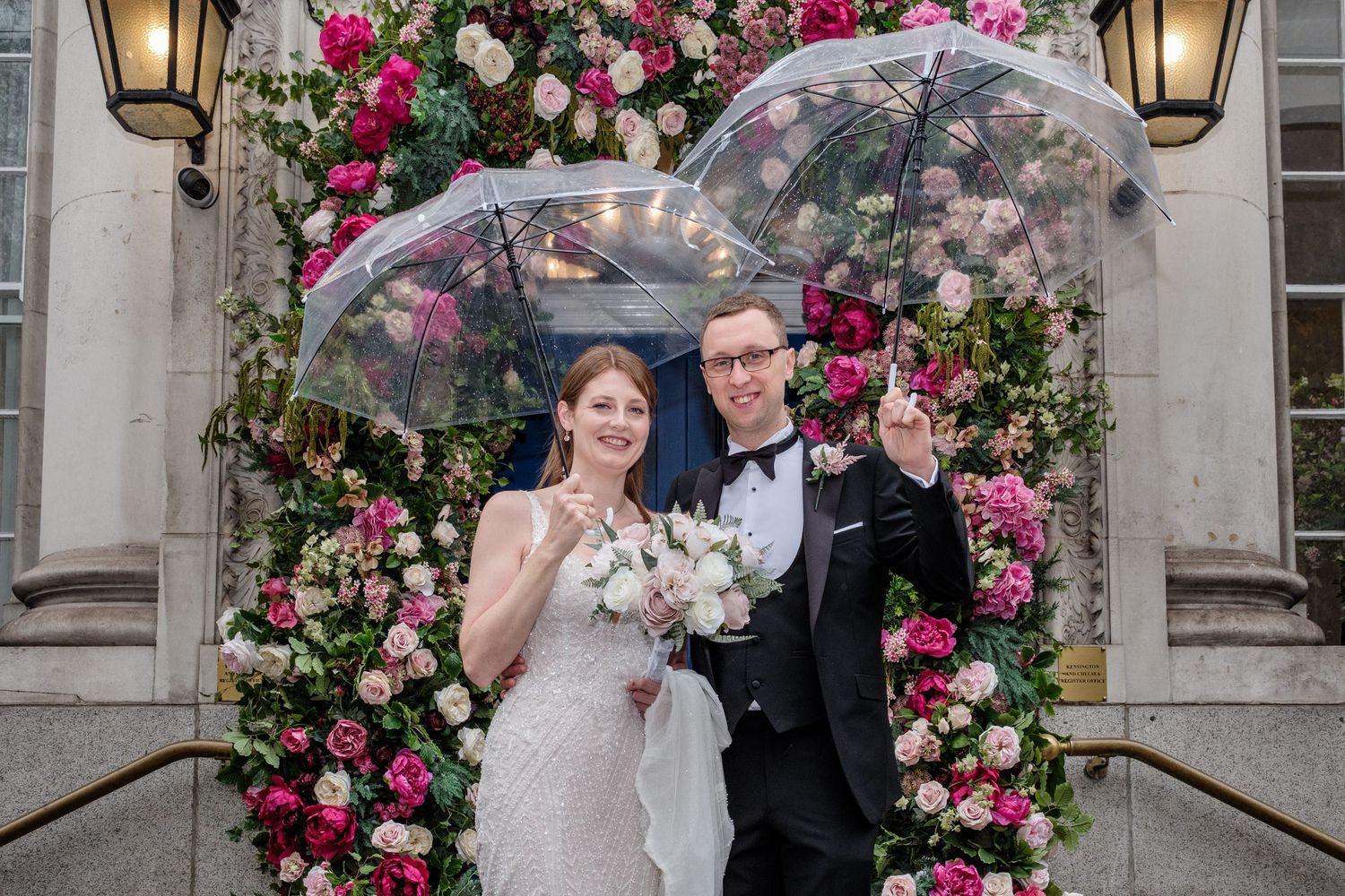 Wedding couple poses under an elegant floral arch with pink and white roses and decorative lanterns.