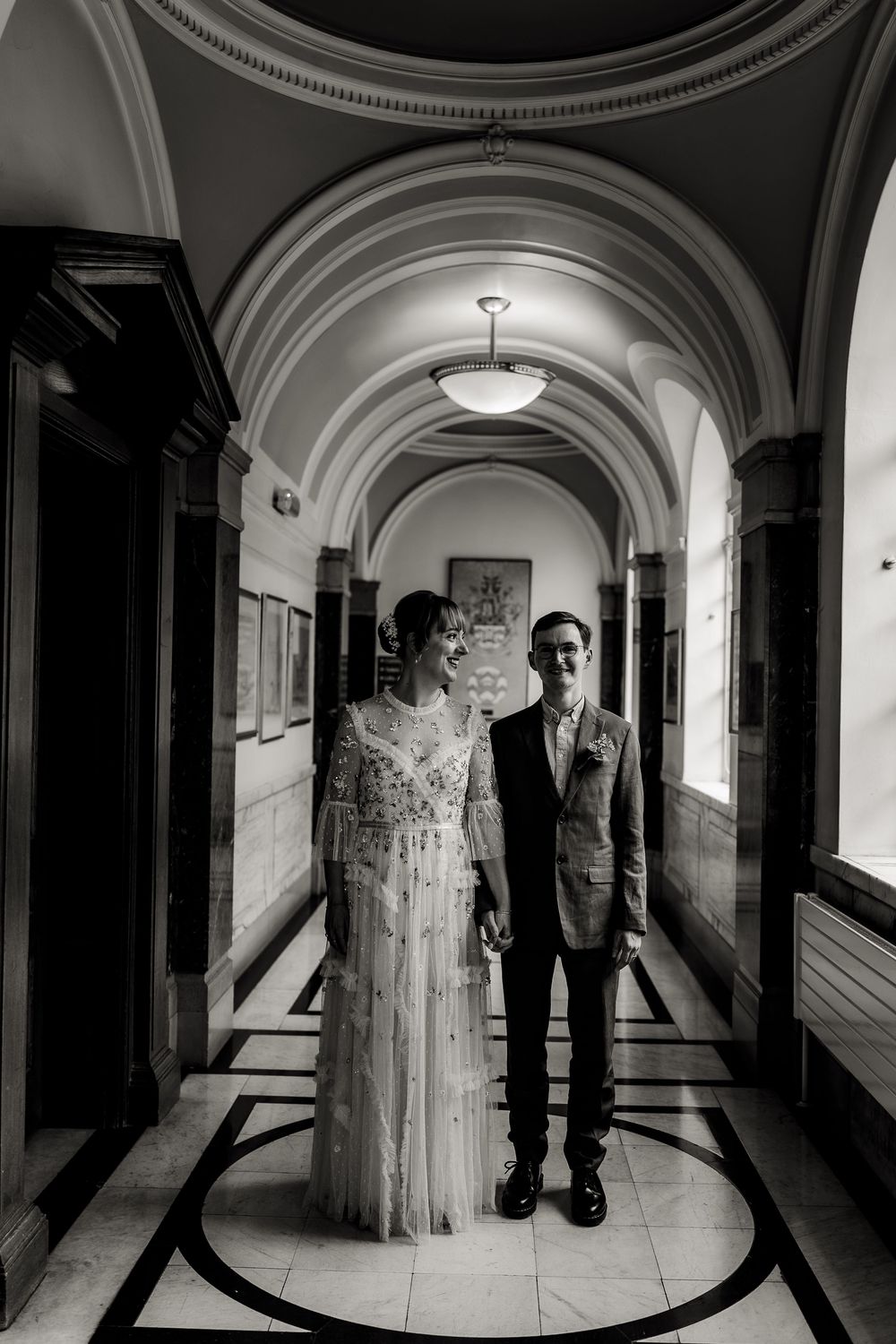 A black and white photograph of a couple standing in an elegant arched hallway with ornate architectural details.