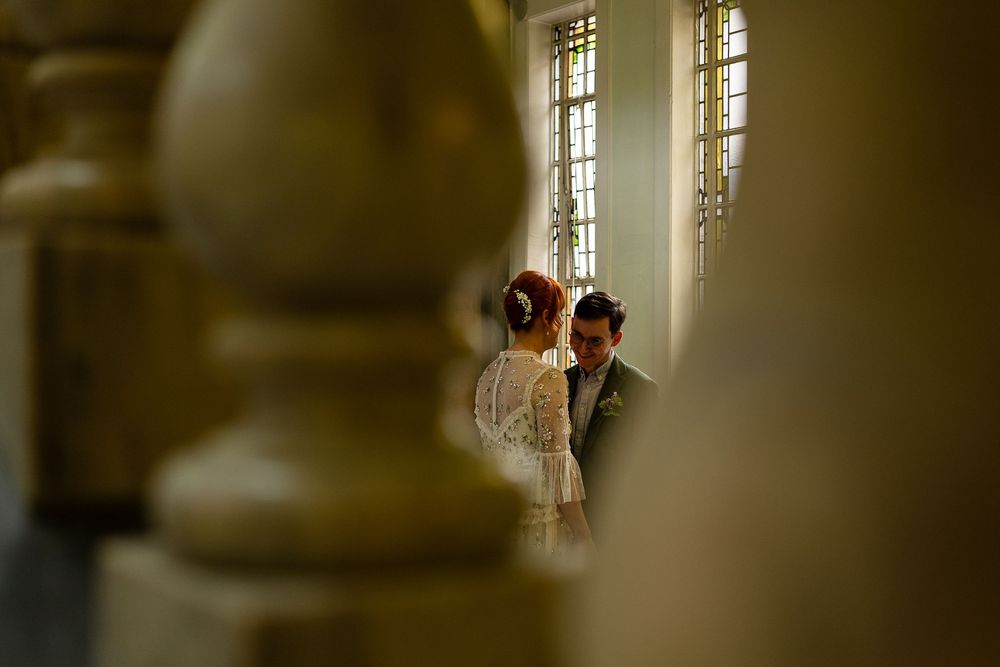 Wedding couple seen through blurred foreground elements in a sunlit room with tall windows.