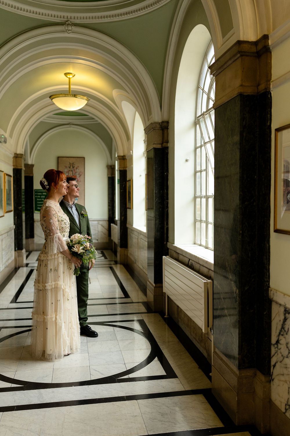 A wedding couple stands in a grand hallway with arched ceilings, marble floors and elegant architectural details.