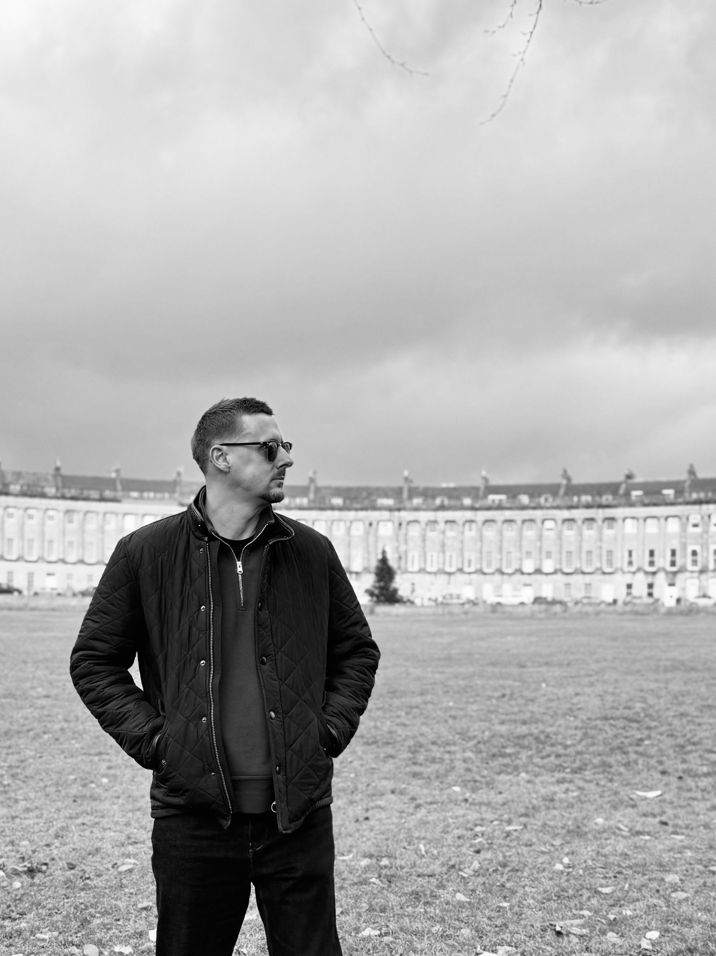 Black and white photo of someone in sunglasses and jacket standing in front of the historic Royal Crescent in Bath, England.