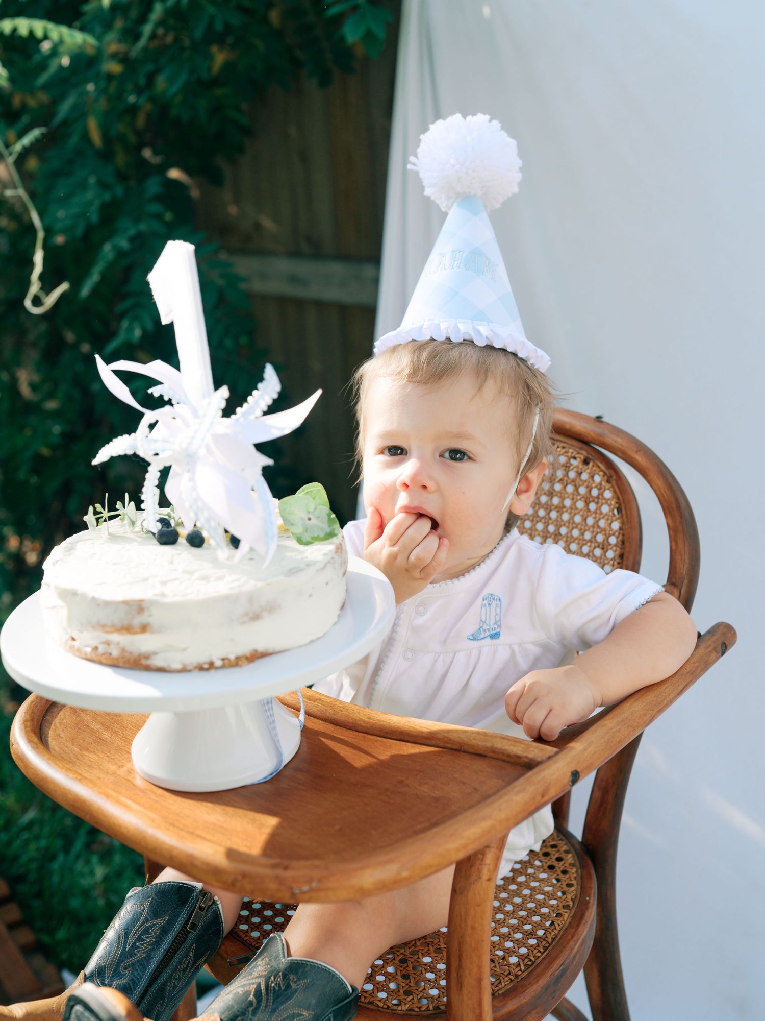 A baby in a wooden high chair reaching for a small white birthday cake with decorative topper on the tray.