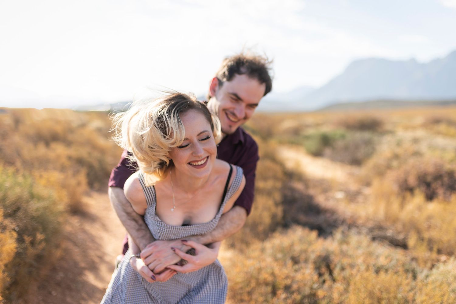 A couple embraces and laughs together on a hiking trail surrounded by desert brush and mountains in the distance.