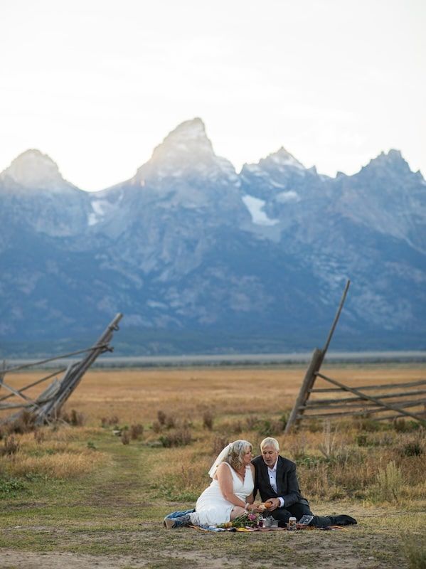 A couple sits on a blanket in front of the majestic Grand Teton mountain range during golden hour.
