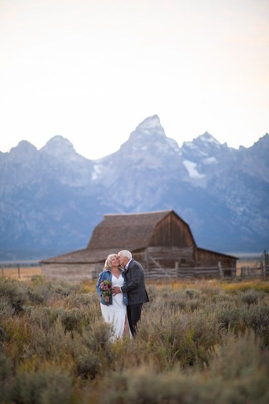 A rustic barn stands beneath dramatic Grand Teton mountains as a couple embraces in the golden autumn field.