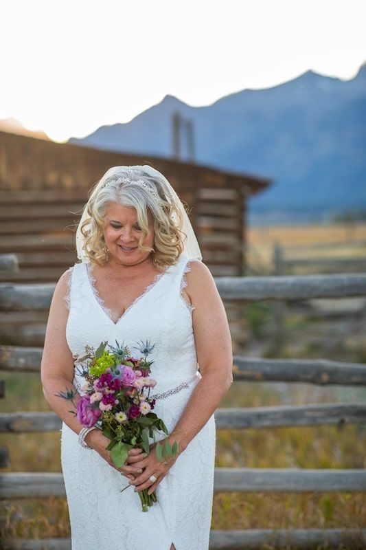 Bride in white lace dress holding vibrant purple and pink floral bouquet against rustic wooden fence at sunset.