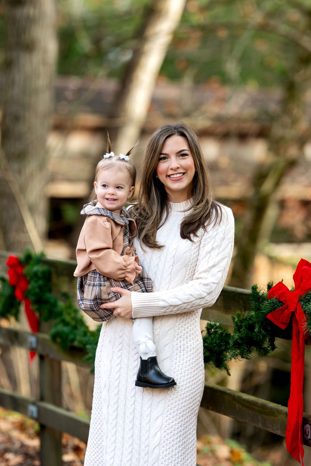 Smiling mother in white knit dress holds toddler in plaid outfit near Christmas-decorated fence.