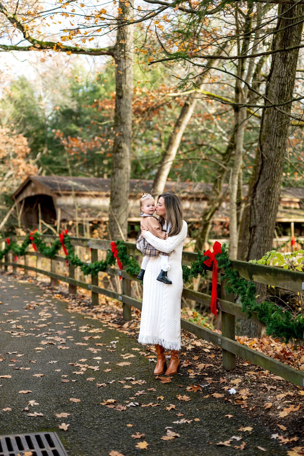 Mother in white dress kisses toddler girl near Christmas-decorated fence and covered bridge in autumn.