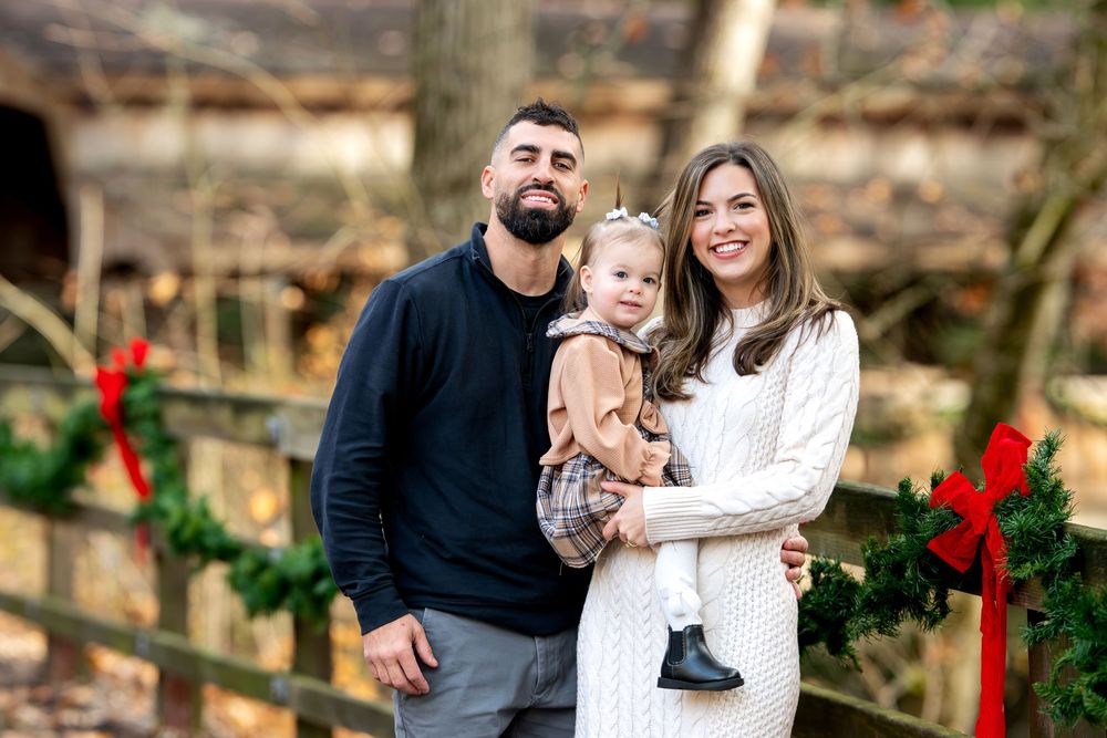 Family of three poses together near Christmas garland and red bows on wooden fence outdoors.