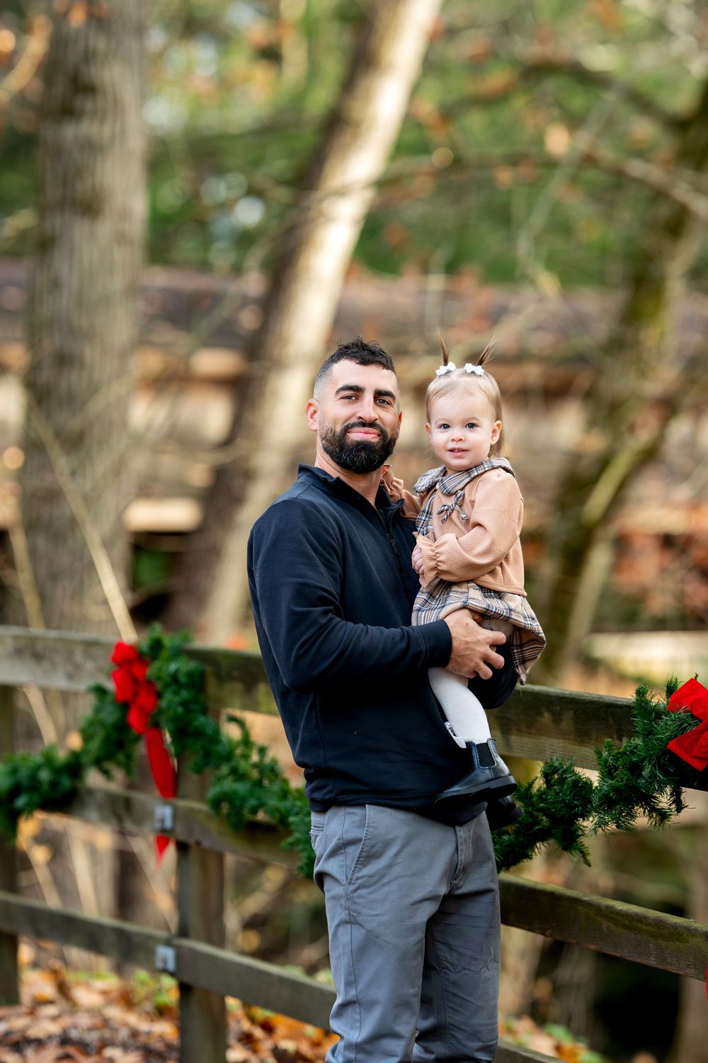 Bearded father holding smiling toddler girl in plaid dress by Christmas decorated wooden fence.