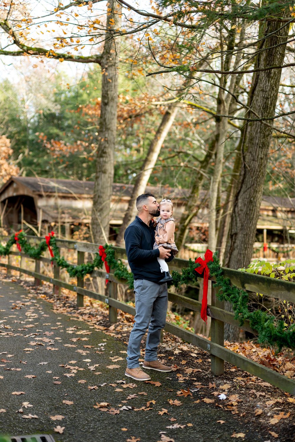 Father kissing baby girl while standing on leaf-covered path with holiday garland fence backdrop.