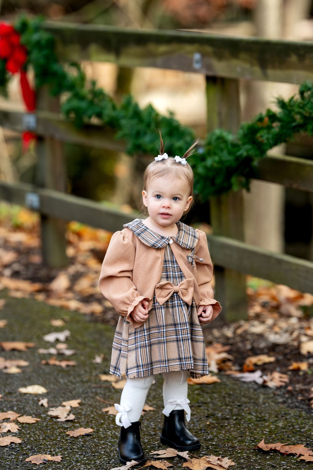 Adorable toddler girl in plaid dress and camel jacket standing on autumn path by garland fence.