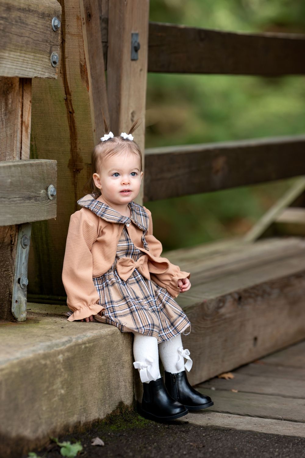 Sweet baby girl in plaid dress and black boots sitting on wooden bridge steps outdoors.