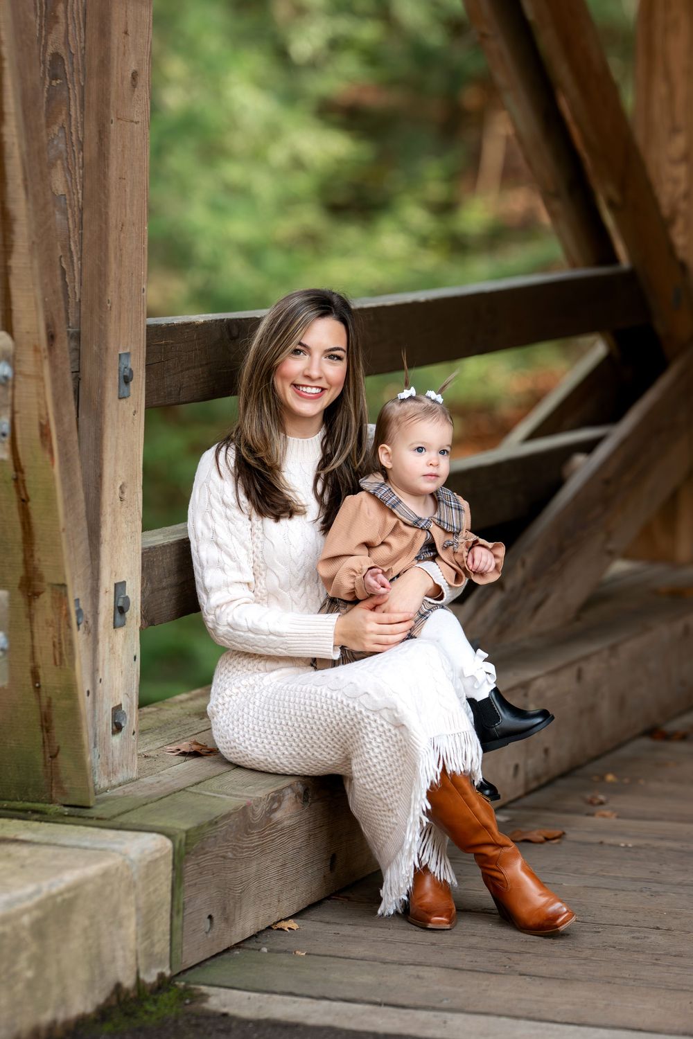 Smiling mother in white dress holding toddler daughter on wooden covered bridge steps in forest.