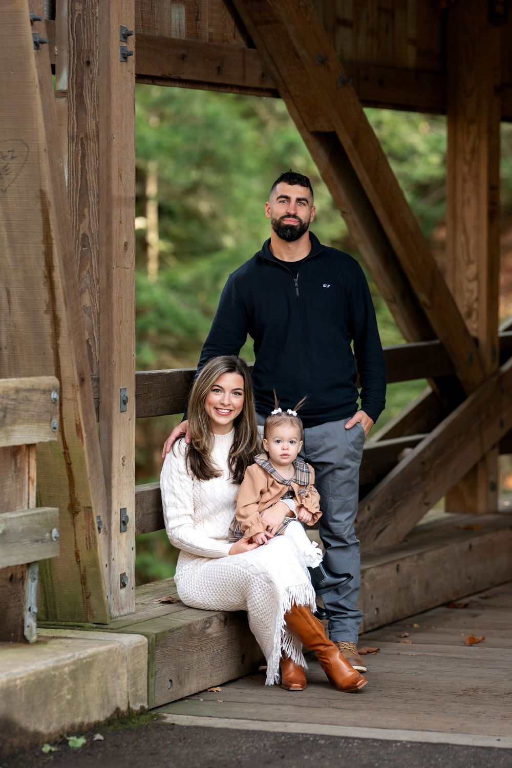 Family portrait of parents with toddler daughter posing inside rustic wooden covered bridge.