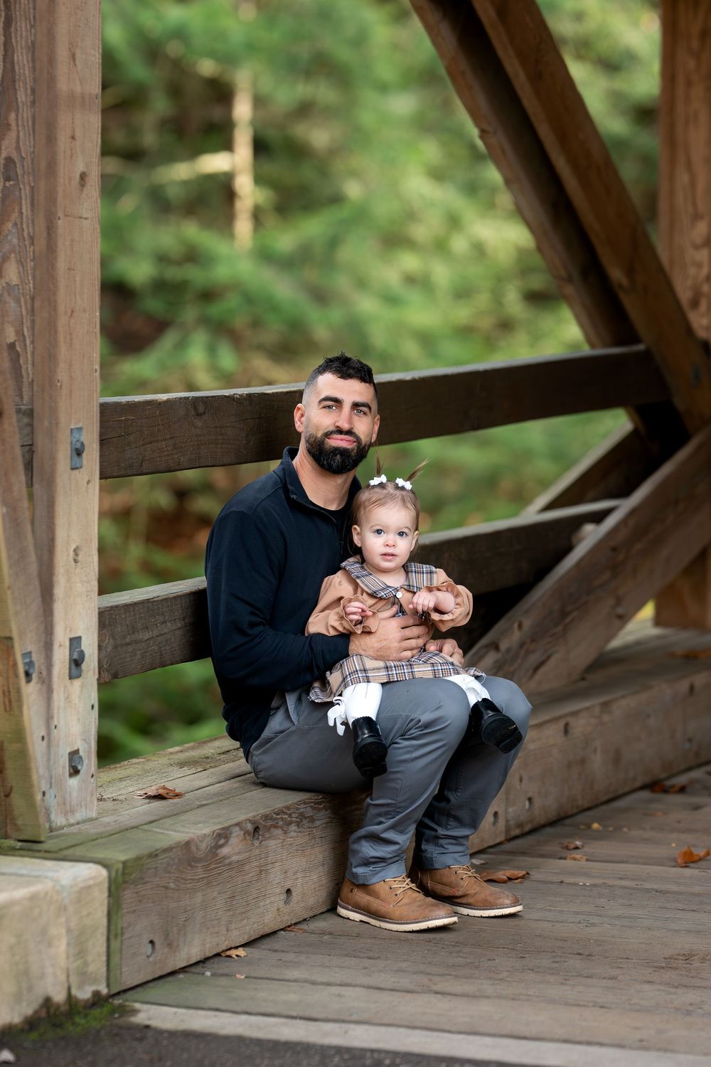 Father sitting on covered bridge steps holding young daughter in plaid dress in green forest.