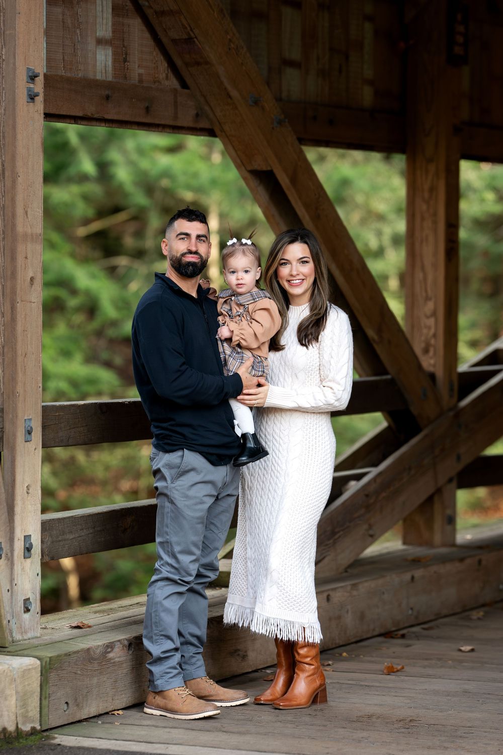 Family portrait of parents holding toddler on wooden covered bridge surrounded by forest.