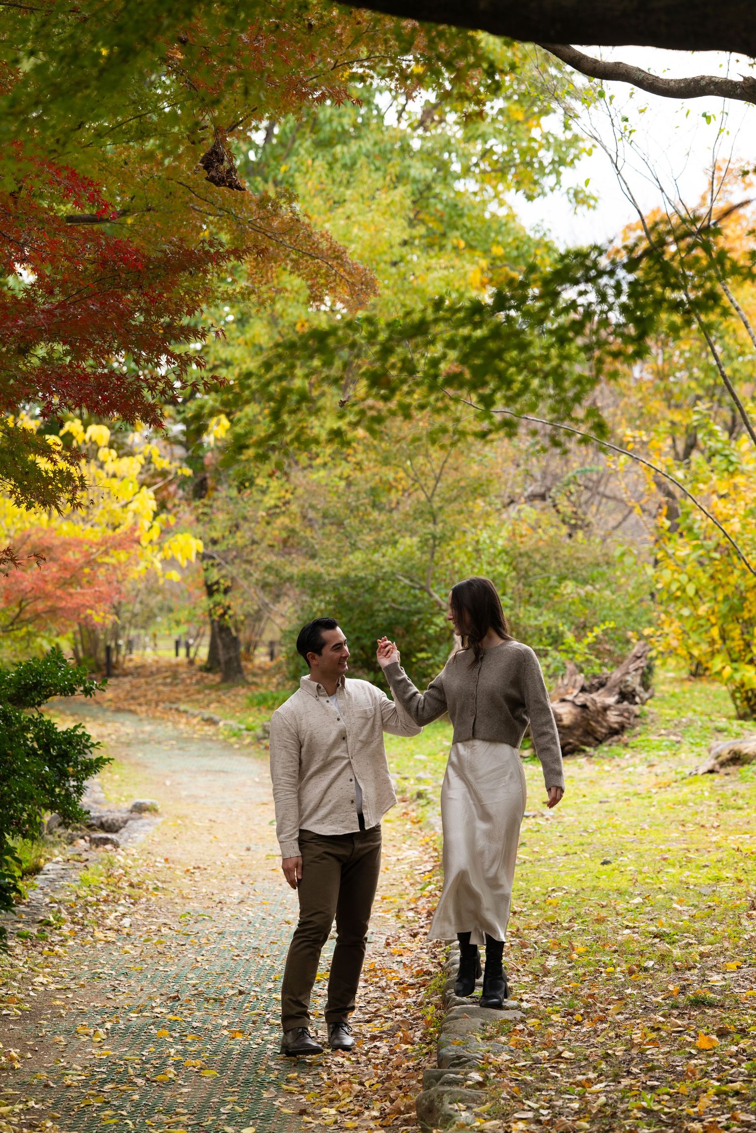 Man spinning woman on a leaf-covered garden path framed by colorful autumn trees.