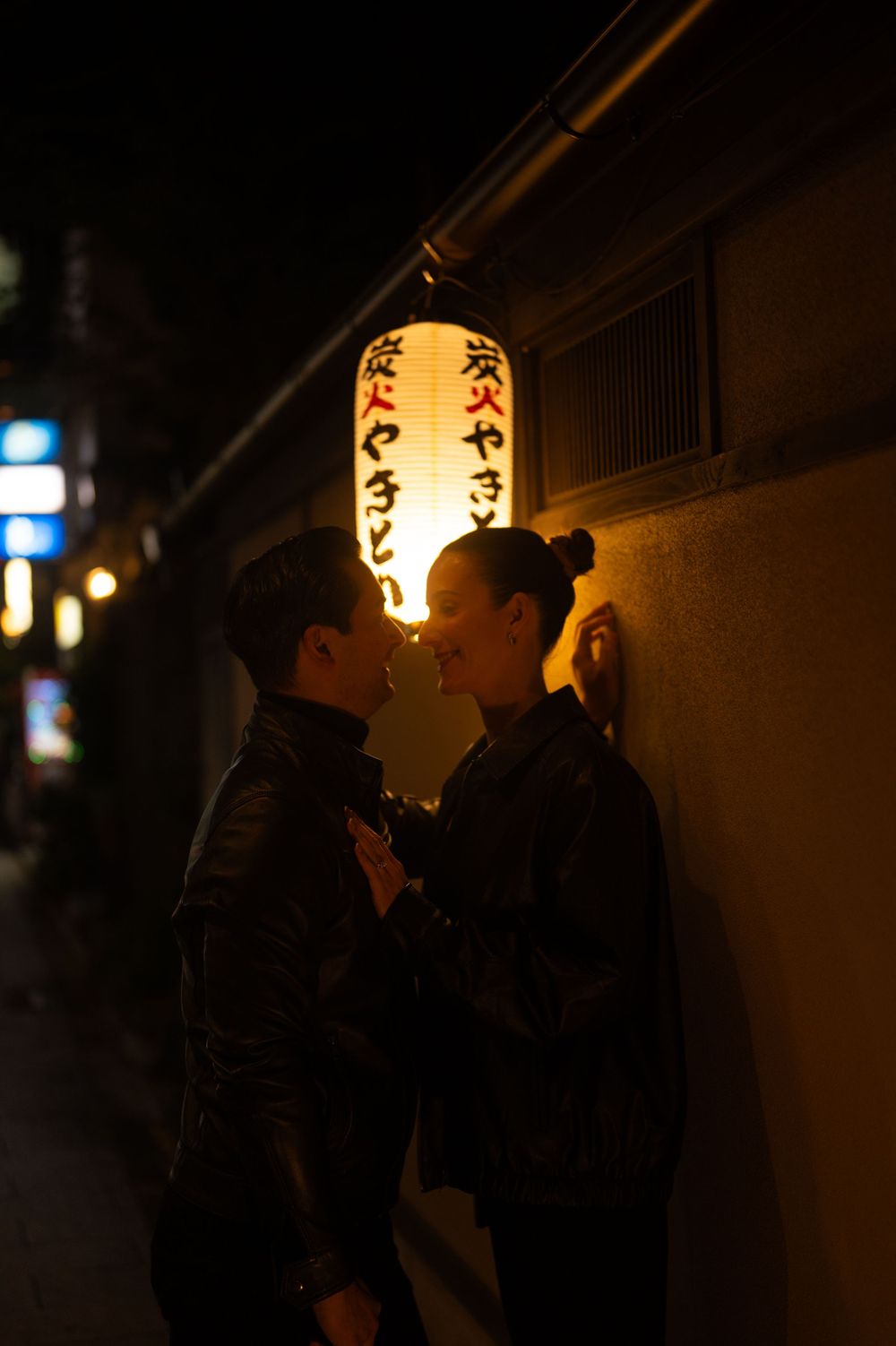 Romantic couple leaning close beneath a glowing Japanese lantern on a dark city street at night.