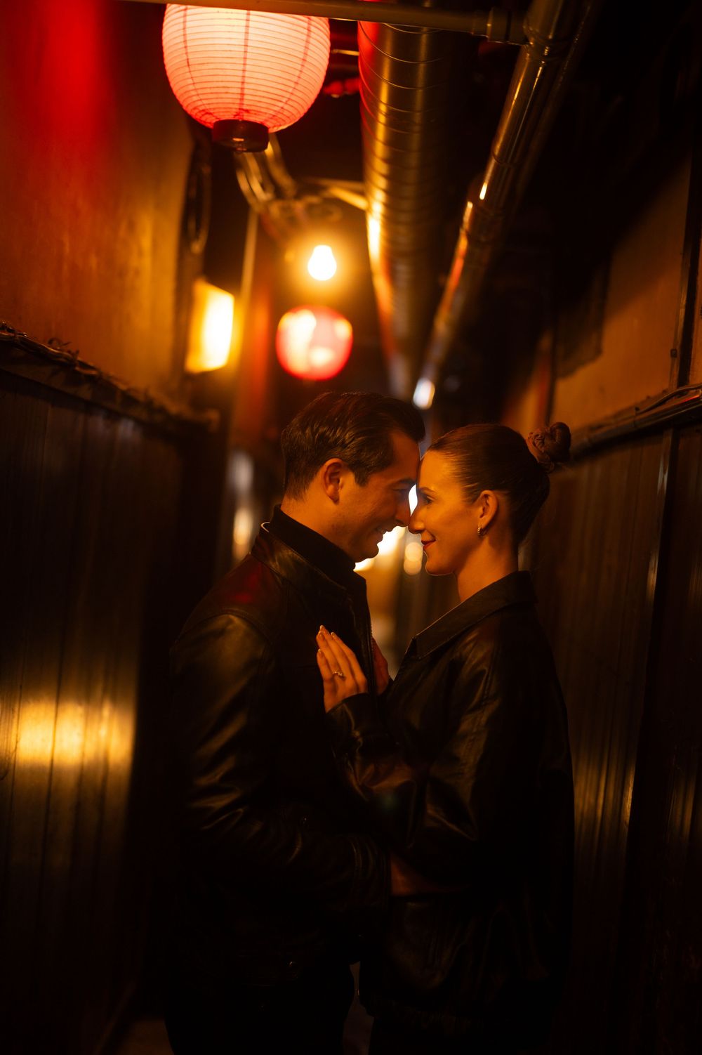 Couple smiling and embracing in a narrow Japanese alley glowing with red and gold lanterns.