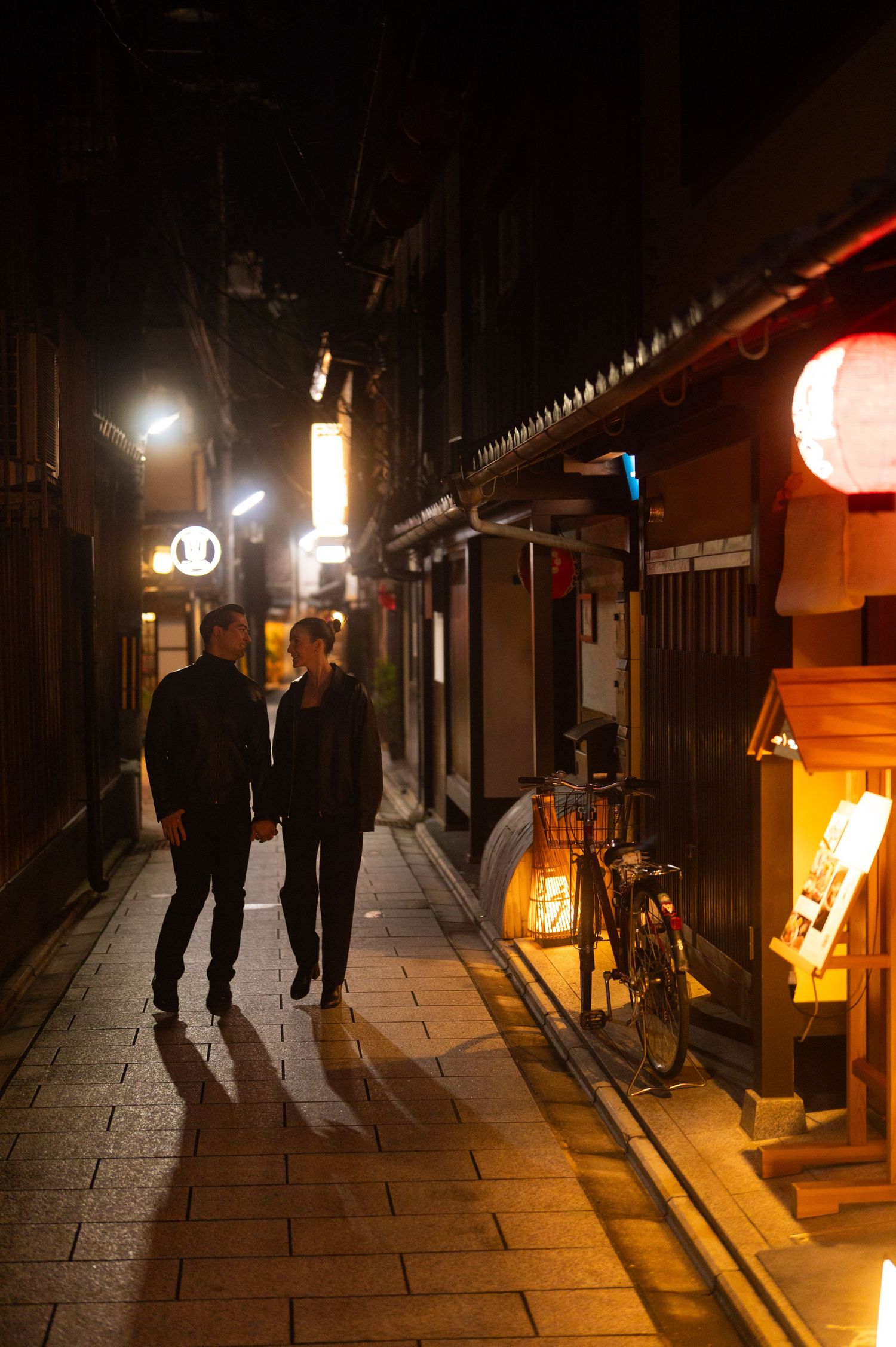 Couple walking hand in hand through a glowing lantern-lit alley in Kyoto at night.