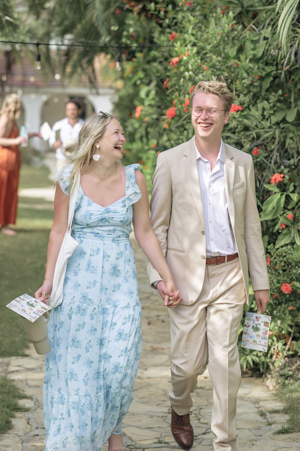 A couple laughs together while walking along a garden path with red flowers and greenery.