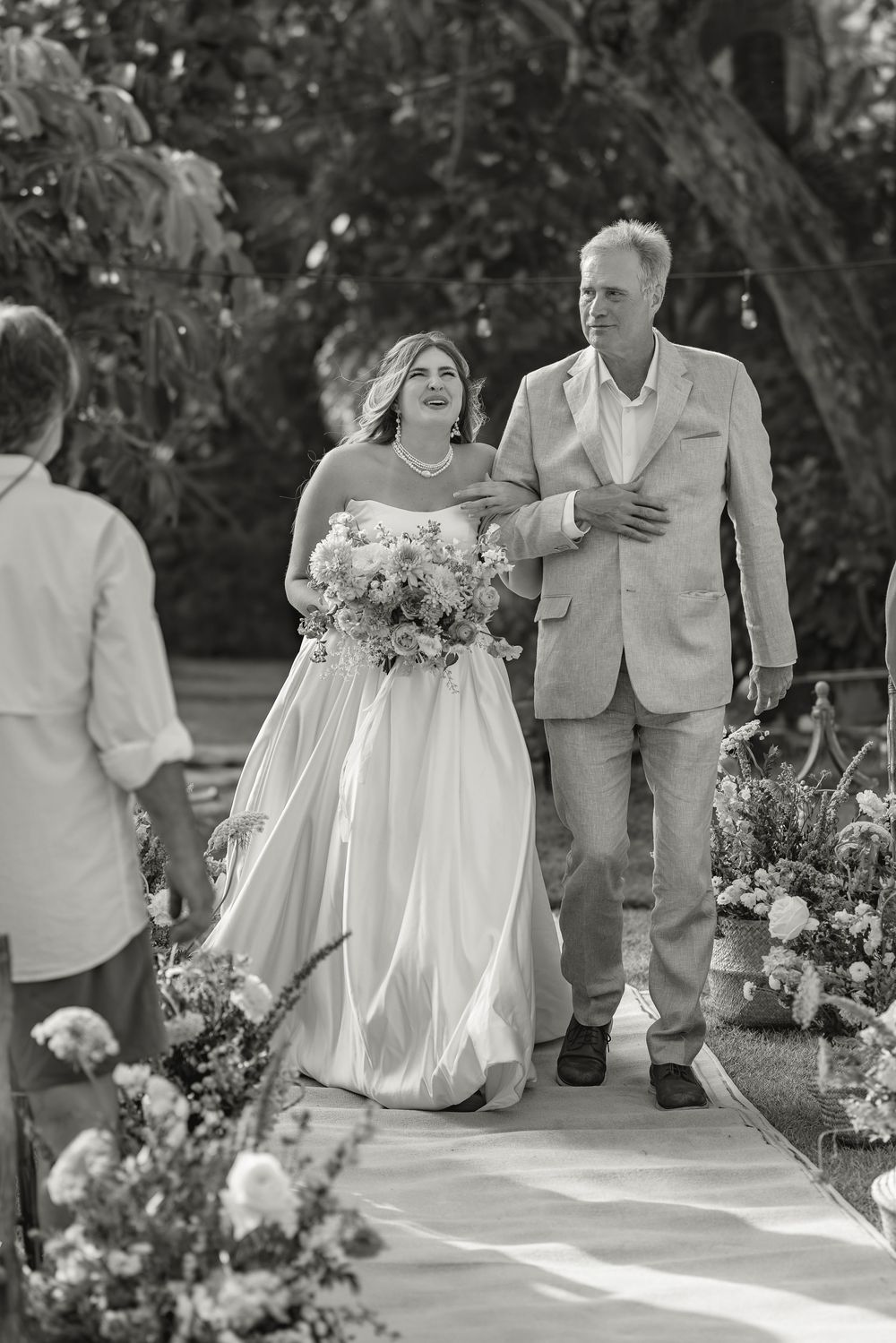 Black and white photo of a wedding ceremony with the bride in a flowing dress walking down a flower-lined path.