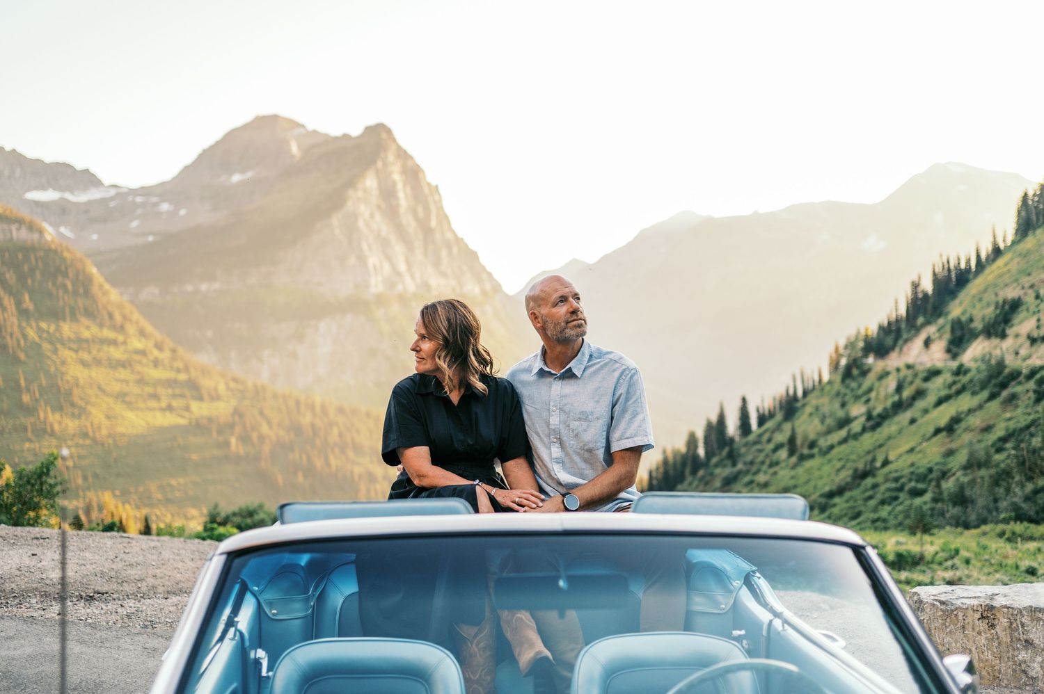 A couple sits on their blue car while enjoying a scenic mountain view during golden hour sunset.