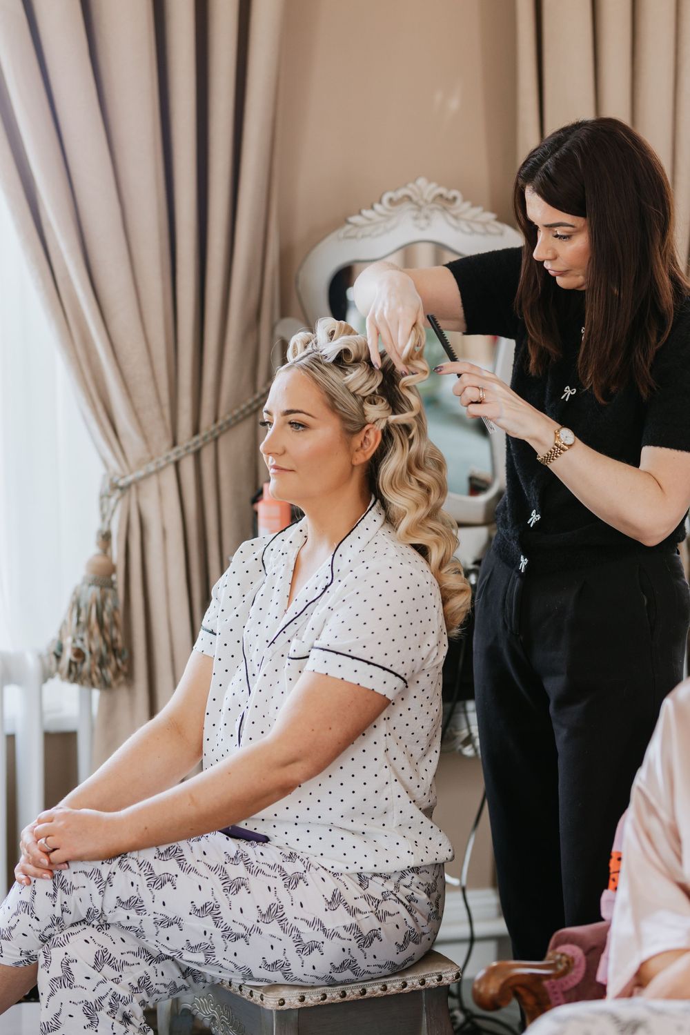 A person in a white polka dot dress sits in a chair while getting hair and makeup done before a special event.