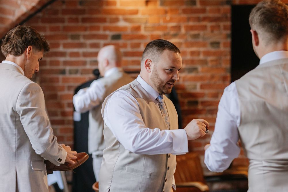 Groomsmen help each other get ready in a rustic brick-walled room while wearing light colored suits.