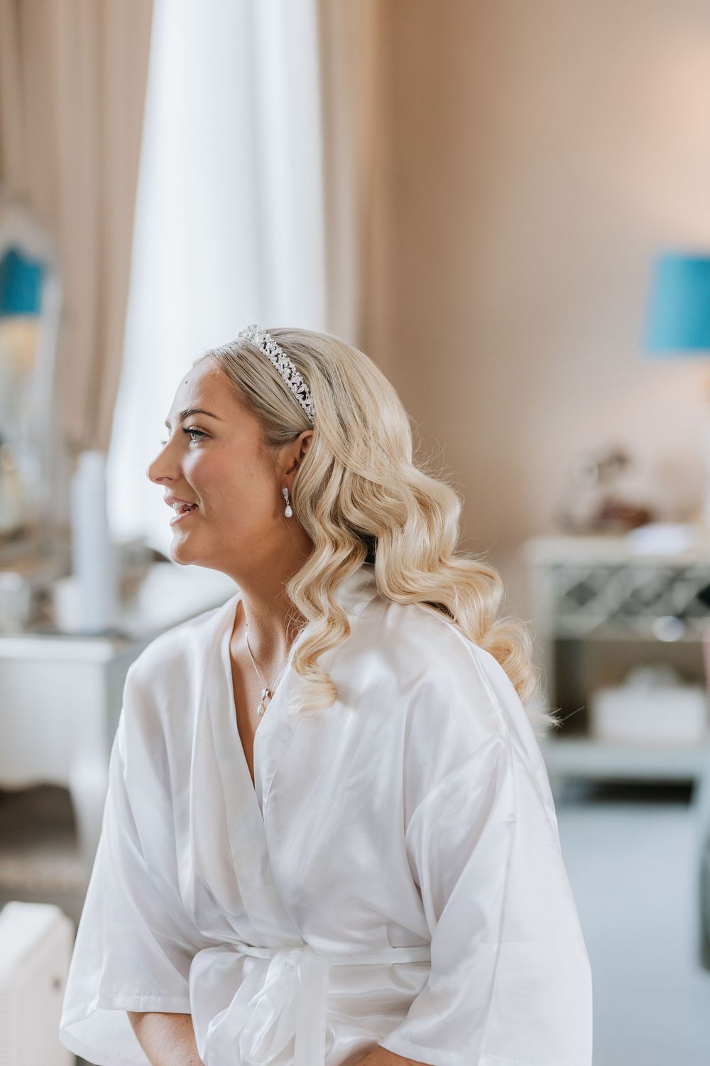 Woman in white robe smiling and looking to the side while getting ready in bright room.