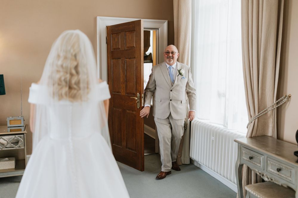 A bride in a flowing white gown and veil stands by a window in an elegant room with natural light streaming in.