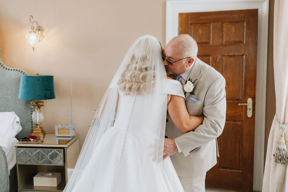 An emotional moment captured at a wedding as two people embrace in a room with a teal lamp and wooden door.