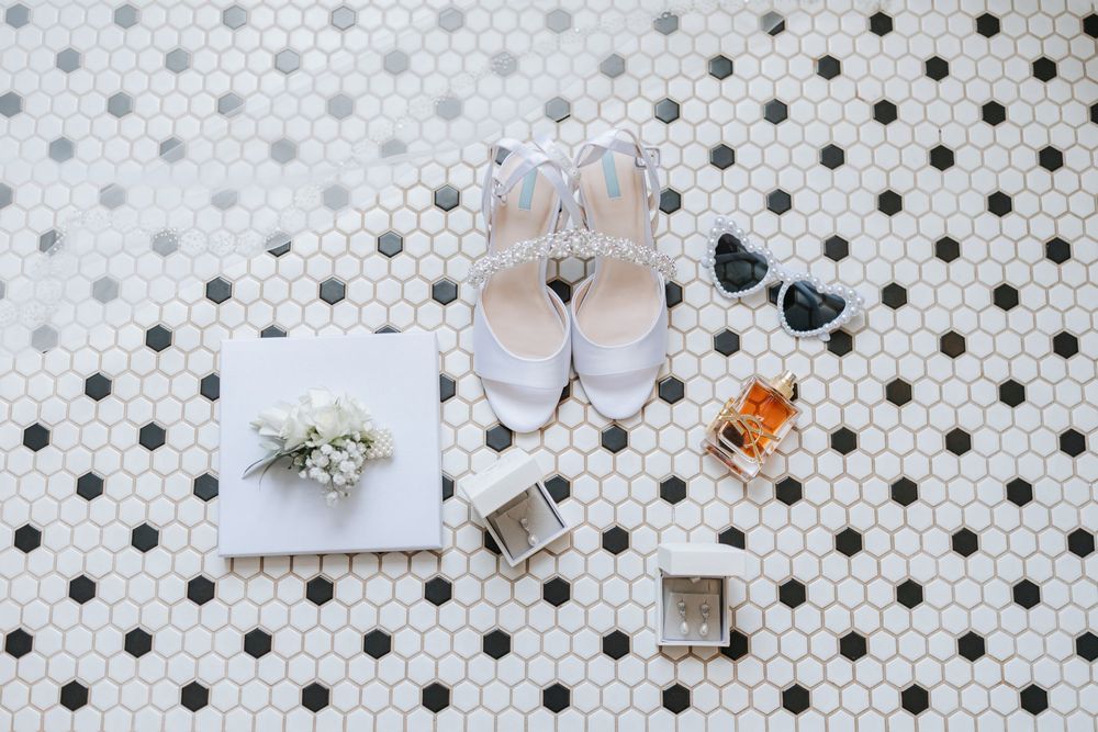 Wedding accessories including blush pink heels, jewelry, and perfume arranged on white polka dot tile floor.