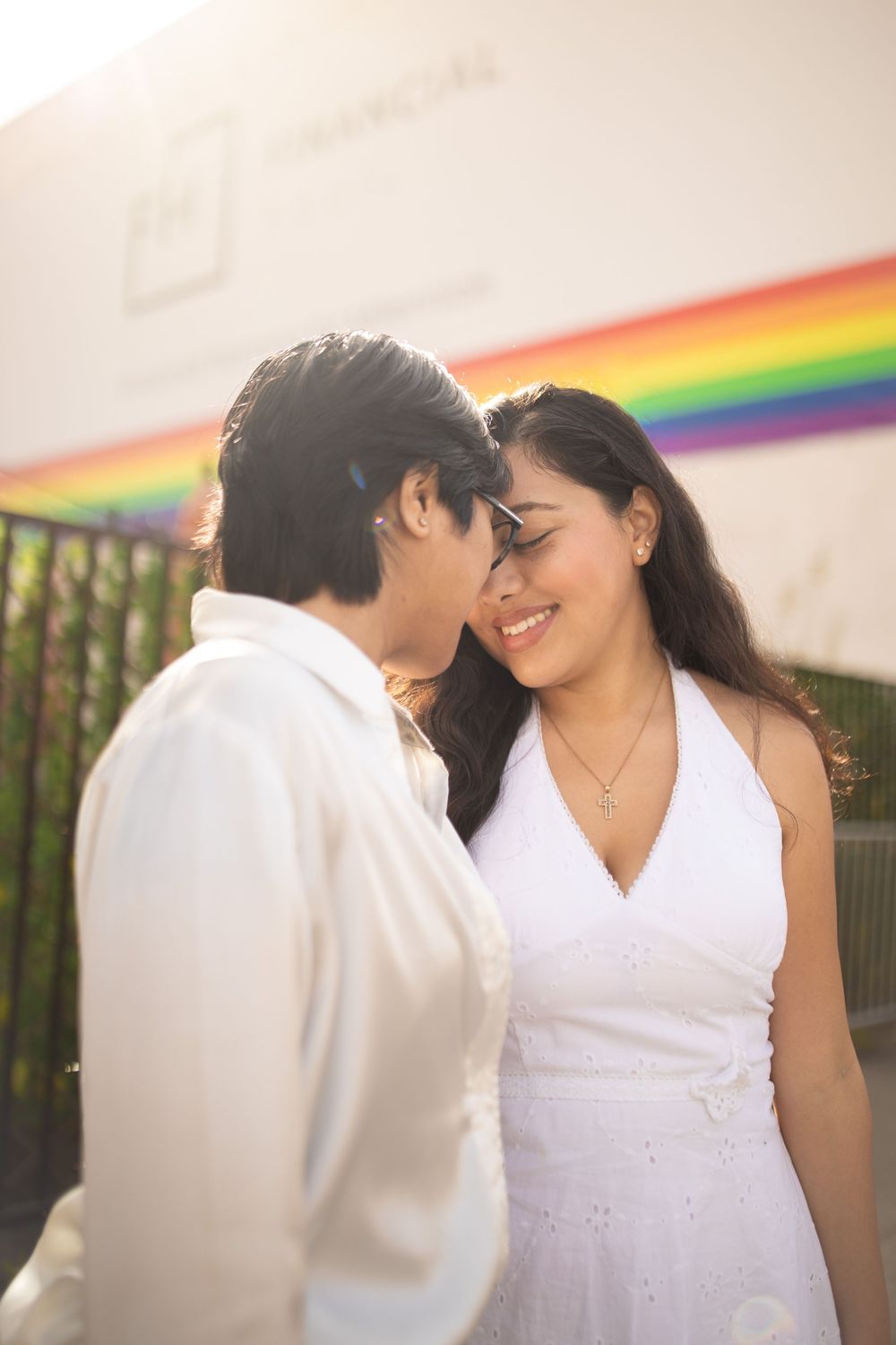 A couple in white outfits poses together against a rainbow backdrop in a series of romantic engagement photos.