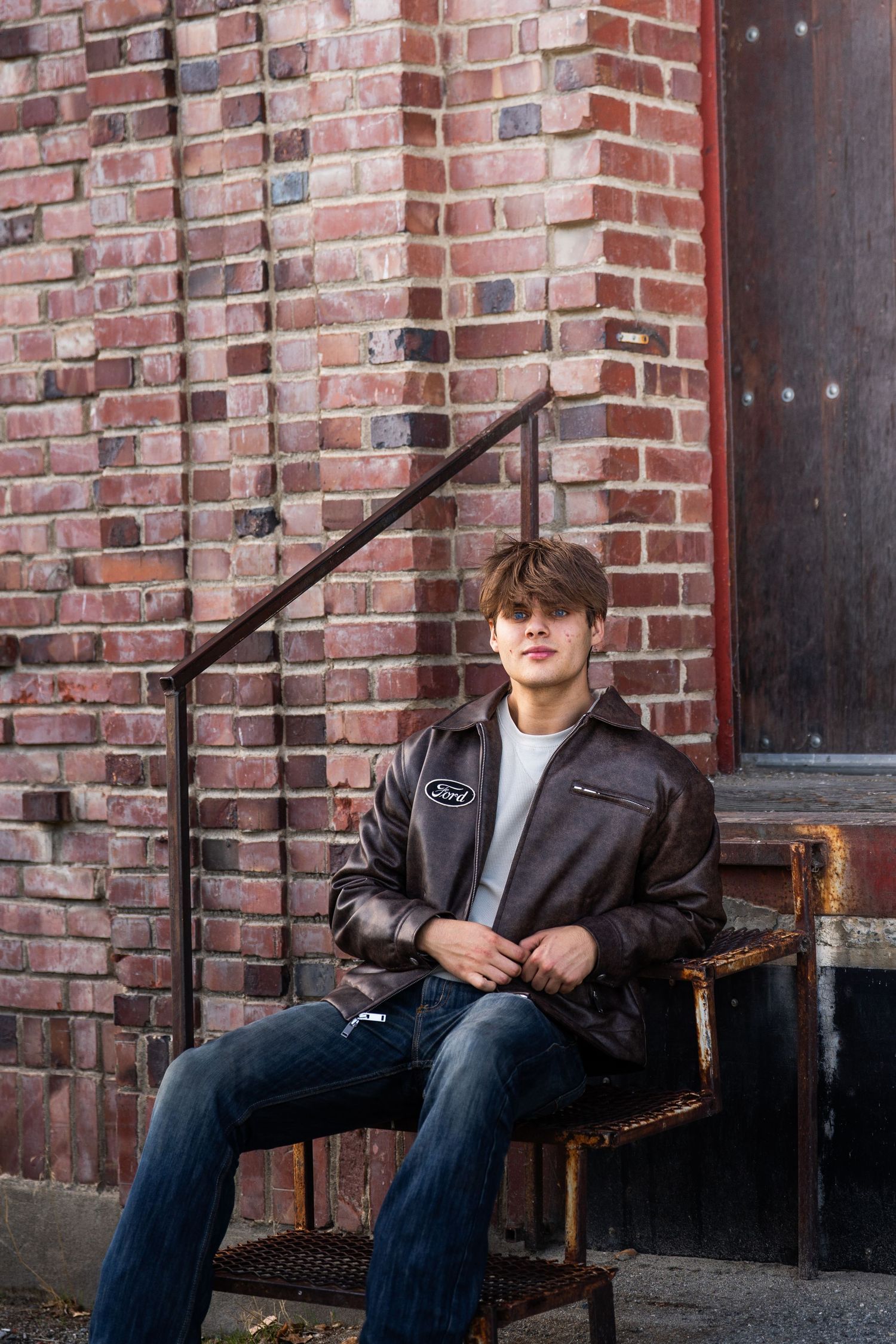 Person sitting on wooden bench against red brick wall wearing dark jacket and jeans.
