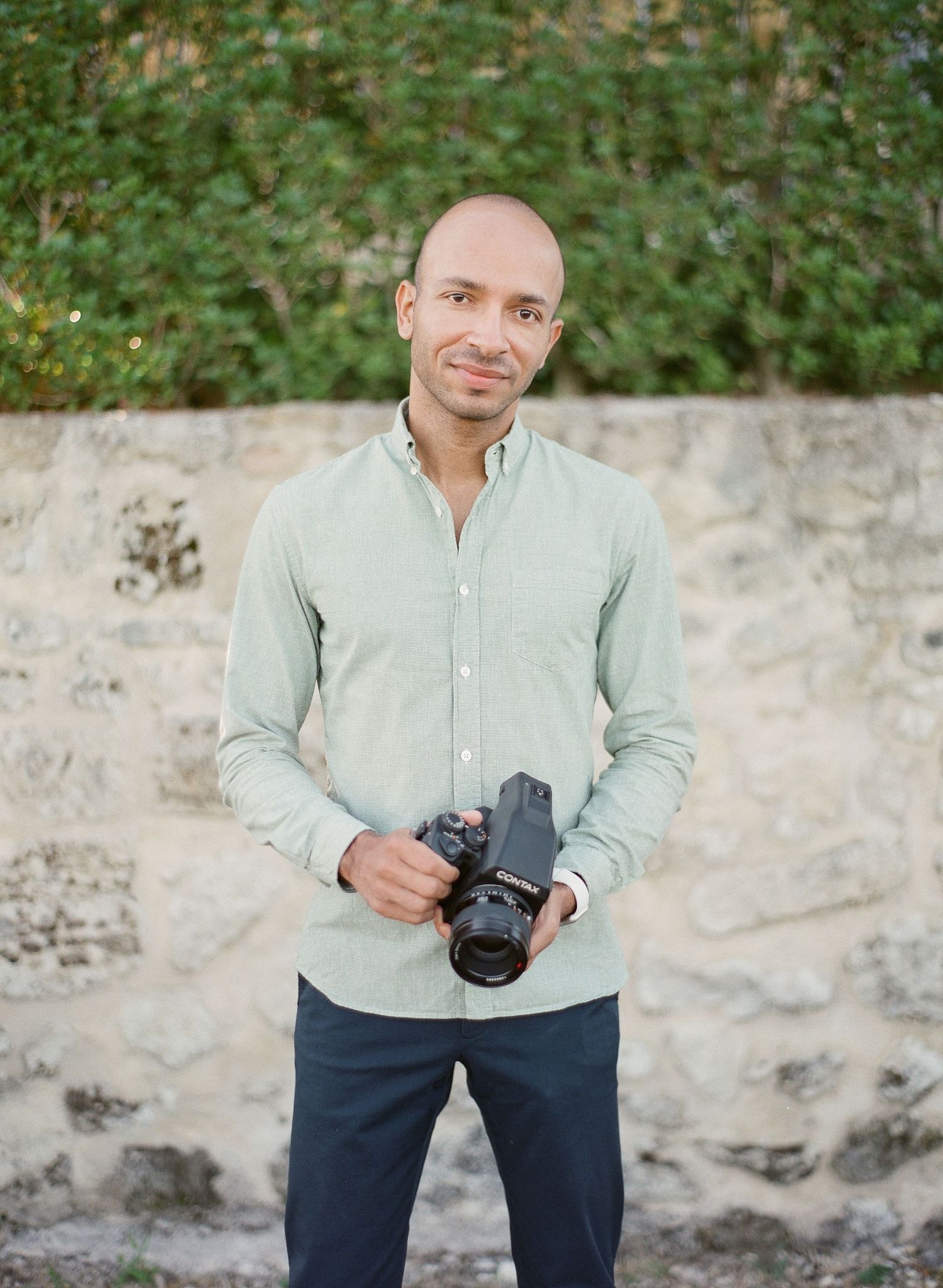 A photographer in a light colored shirt and dark pants holds a camera while standing against a stone wall.