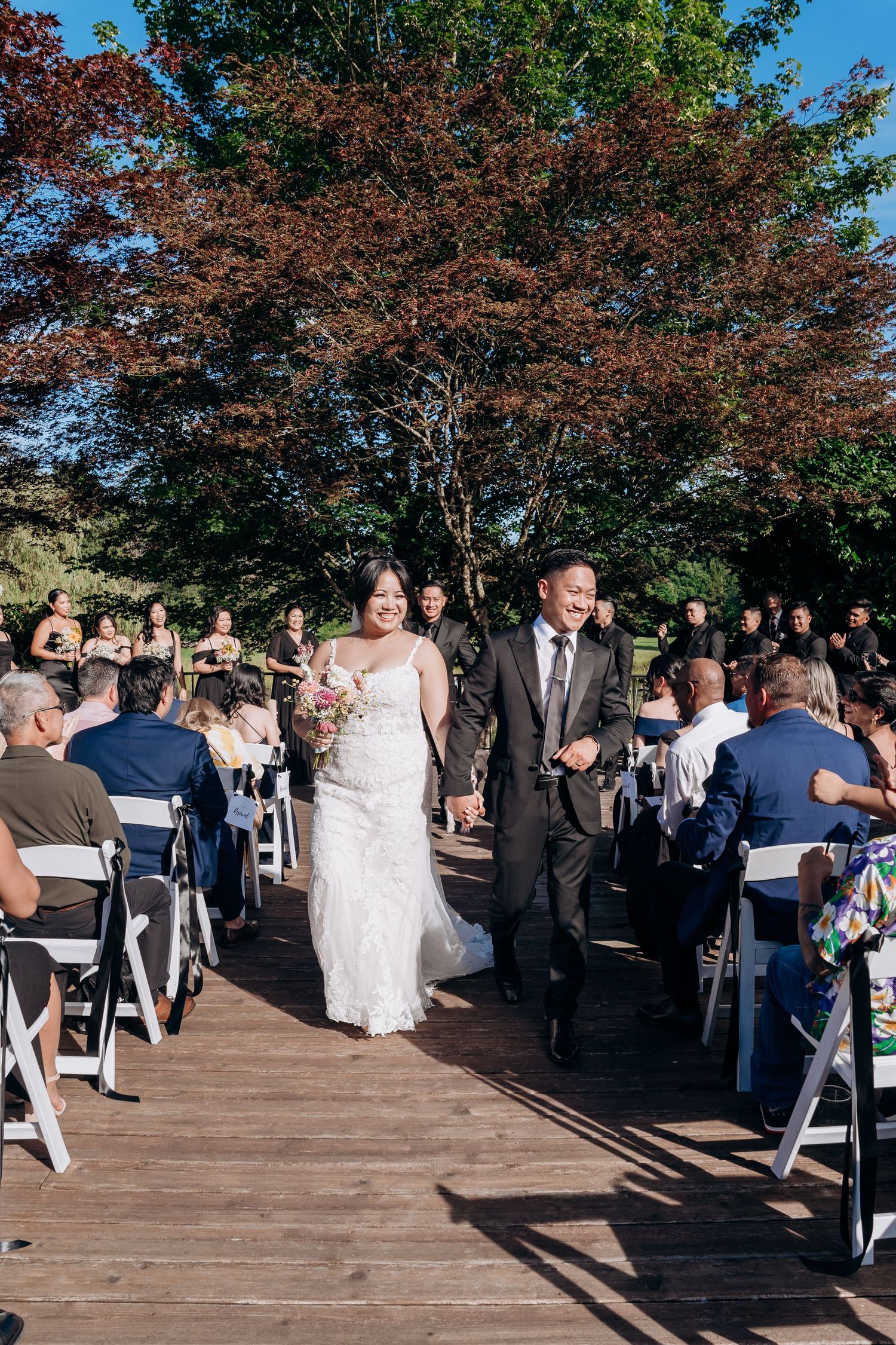 Wedding ceremony guests gather on an outdoor wooden deck surrounded by autumn trees during a late afternoon celebration.