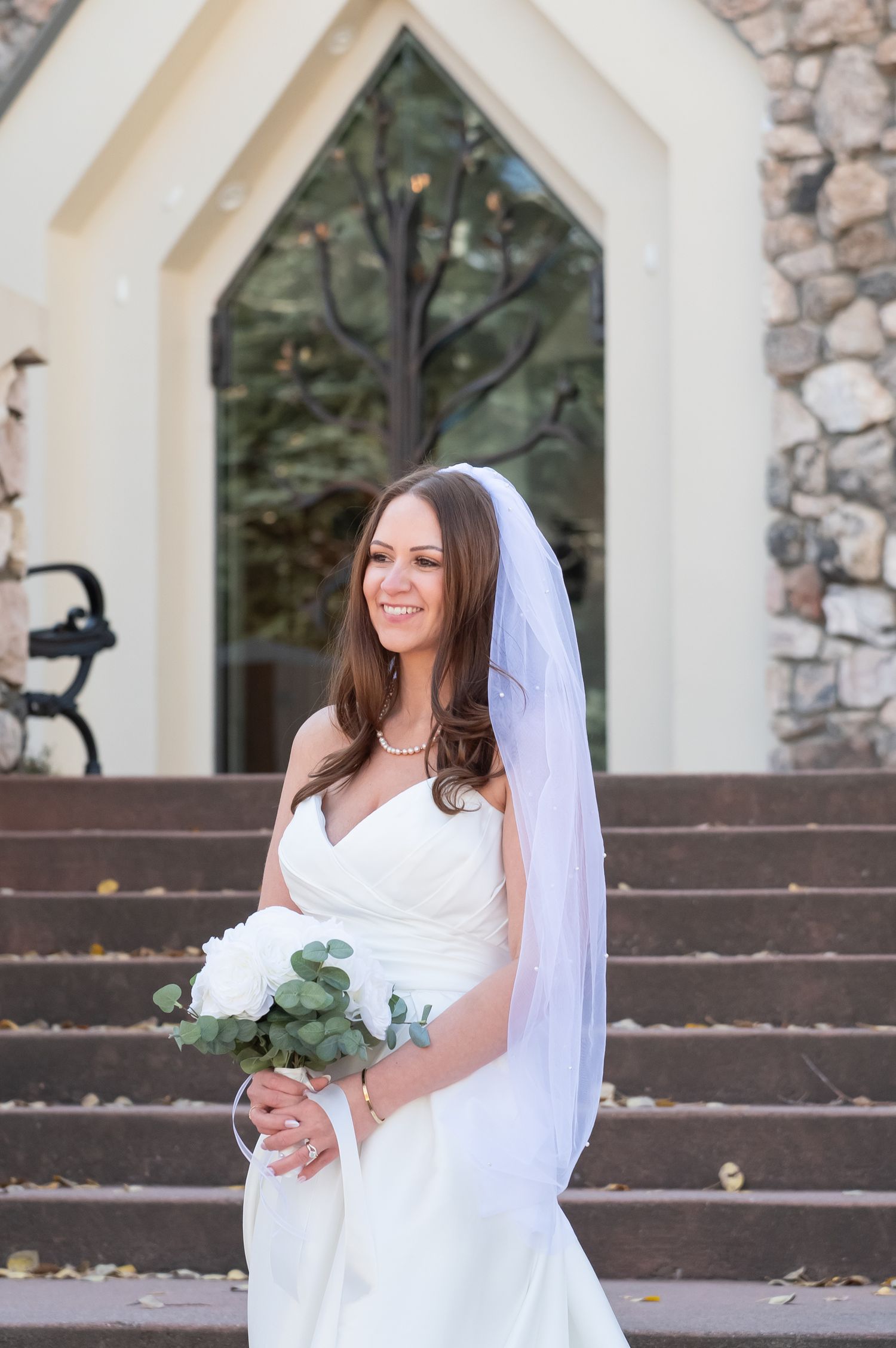 Bride in a white wedding dress and veil smiles while posing on stone steps outside The Beaver Creek Chapel