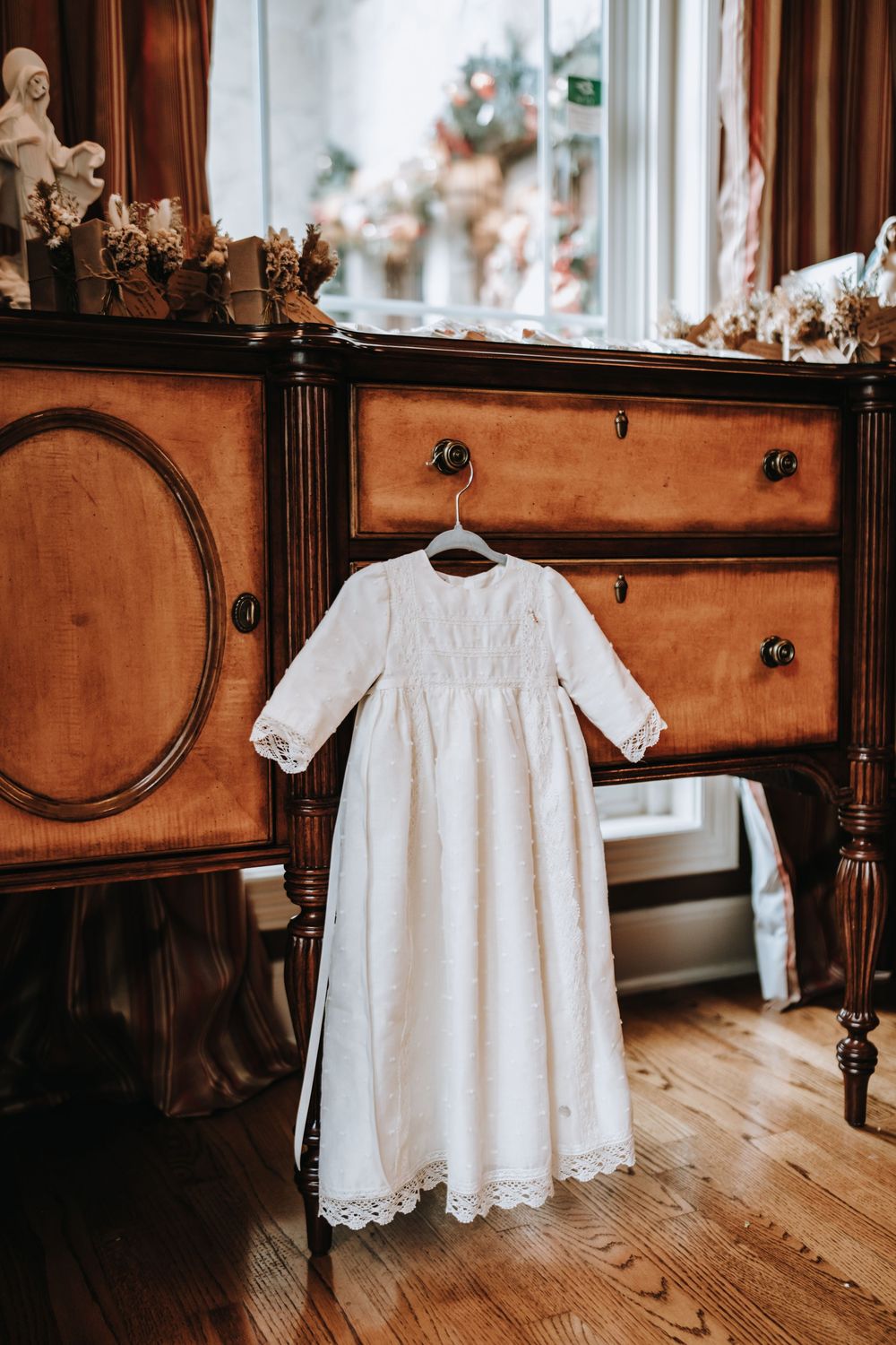 Delicate white christening gown with lace trim displayed against wooden furniture in natural light.