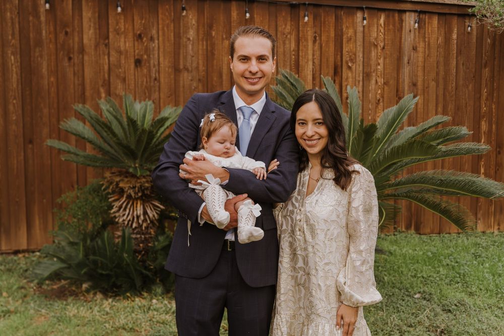 Parents hold infant in formal attire against wooden fence and tropical landscaping backdrop.