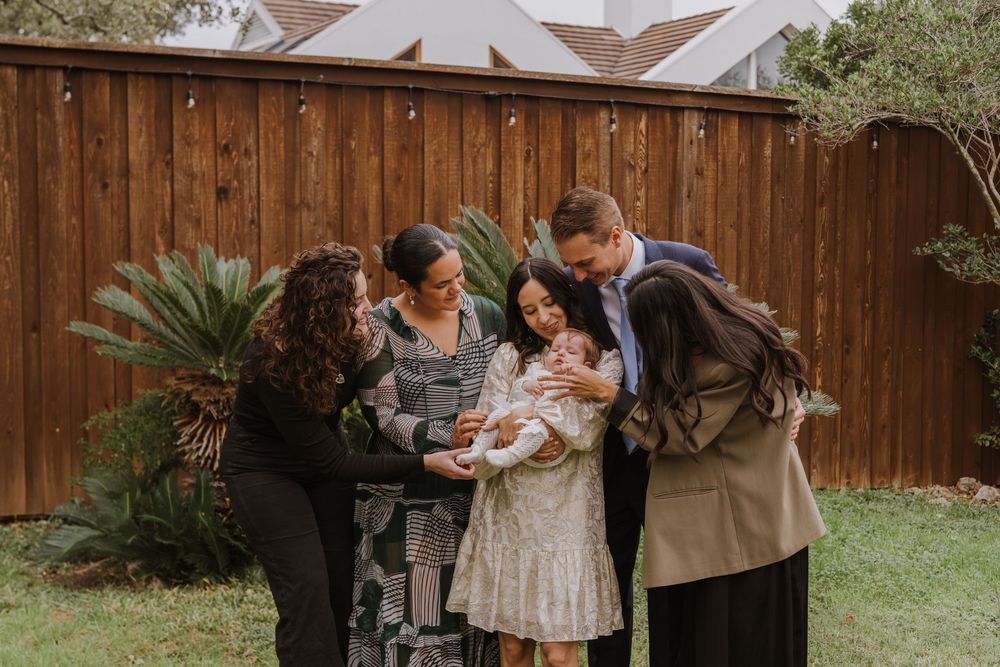Small group gathers closely around baby in white dress in backyard setting.