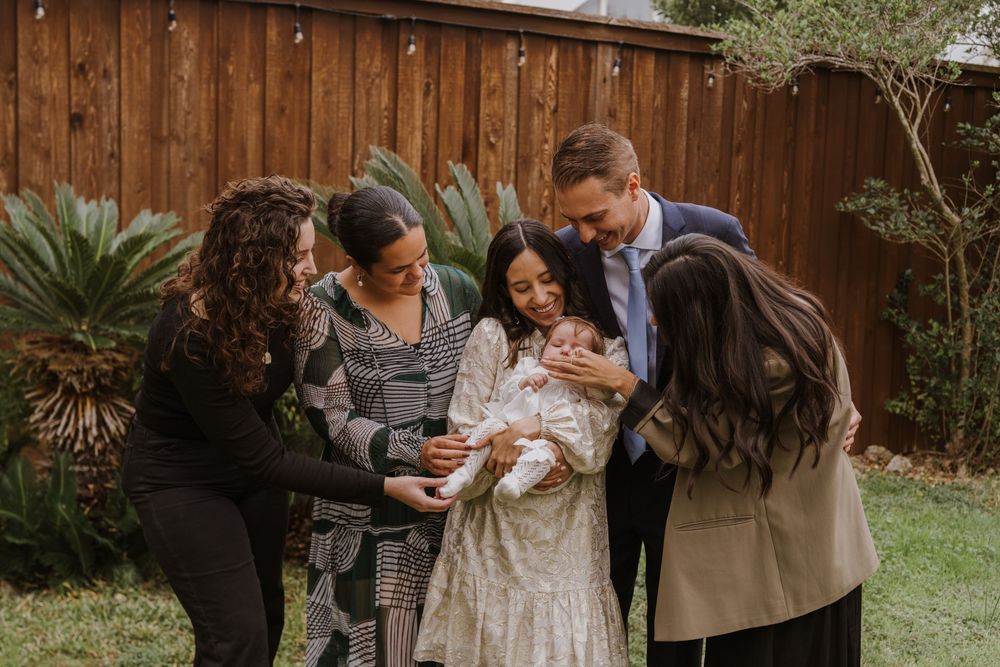 Group huddles together looking at baby in white dress against wooden fence background.