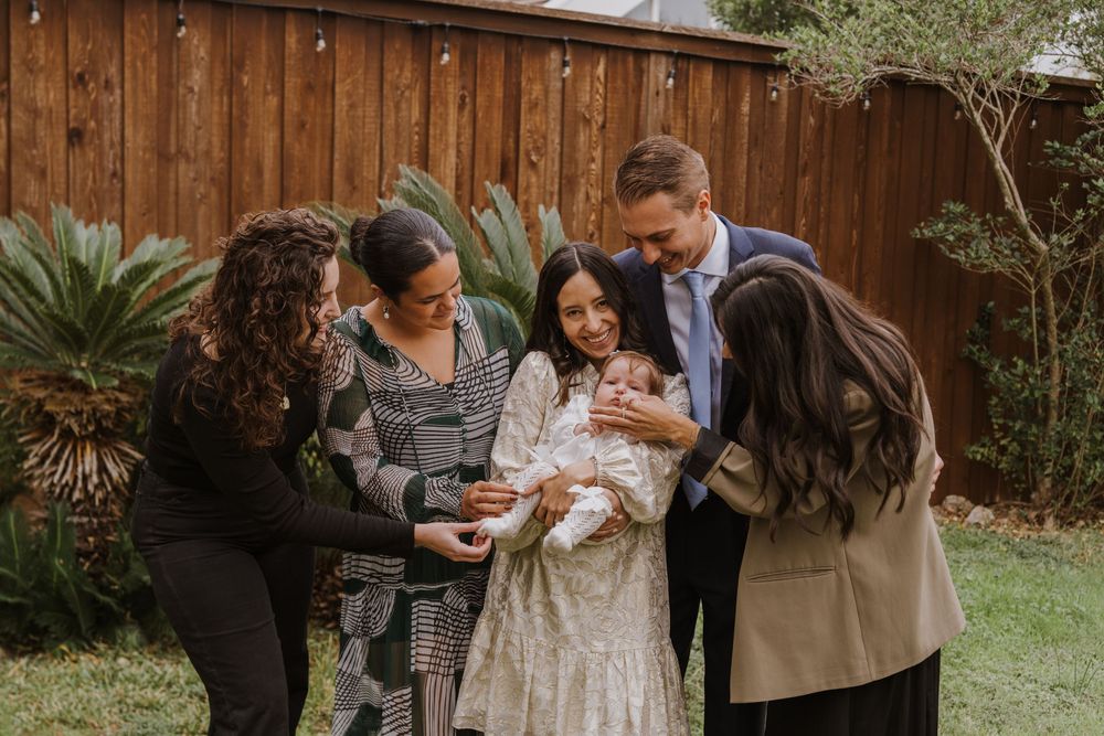 Family members gather closely around a baby in white dress against wooden fence backdrop.